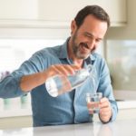 Middle age man drinking a glass of water with a happy face standing and smiling